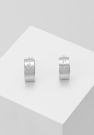 Boucles d'oreilles créoles en argent avec une finition lisse, comportant un logo de marque gravé d'un côté. Présentées sur une surface blanche plate.