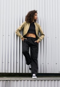 Leichte olivefarbene Bomberjacke über einem schwarzen Cropped-Top, kombiniert mit locker sitzenden schwarzen Cargo-Hosen und schwarzen Sneakers, vor einem grauen Hintergrund.