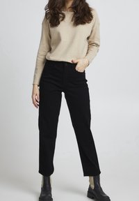 Mujer con suéter de manga larga beige, pantalones altos negros y botas negras, de pie con una mano en el bolsillo frente a un fondo liso.