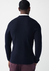 Laivastonsininen neulepaita, jossa on teksturoitu kuvio, resorireunaiset hihat ja helma sekä pyöreä pääntie, puettuna valkoisen paidan päälle.