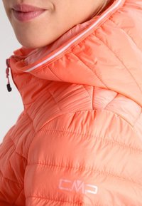 Gros plan sur une personne portant une veste matelassée corail avec une capuche et un logo CMP sur l'épaule.