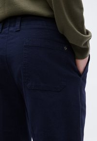 Βαμβακερό παντελόνι σε ναυτικό μπλε με πίσω τσέπη που έχει διακριτική ραφή, συνδυασμένο με ελαιί πράσινο πουλόβερ με υφή.