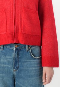 Rode gebreide ritstrui met voorzakken, gedragen met klassieke blauwe denim jeans; hand zichtbaar ontspannen aan de zijkant.