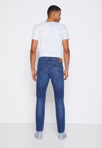 Jeans de mezclilla azul con un corte clásico, que presentan un parche de cuero con el logo de Lee y bolsillos traseros, combinados con una camiseta de tie-dye azul claro.