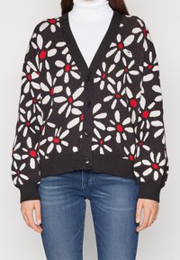 Black cardigan with white and red floral pattern, featuring a V-neck, button front, and ribbed cuffs. Worn over a white turtleneck.