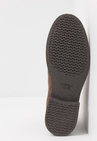 Zapato de ante marrón con una suela de goma texturizada gris que presenta un patrón ondulado, marcado con "GEOX WORLD PATENT" para la marca.