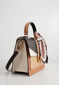 Sac à main beige et noir au design structuré, présentant une texture tissée, une quincaillerie dorée, et un foulard en soie décoratif noué à la poignée.