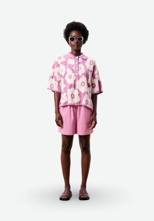Femme portant des lunettes de soleil à monture blanche, un polo à manches courtes rose à fleurs, un short rose assorti et des sandales slides violettes, debout sur un fond blanc.