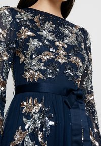 Marinblå klänning med långa, genomskinliga ärmar, prydd med intrikata paljettblommor broderade i silver och brons, samt ett tygbälte i midjan.