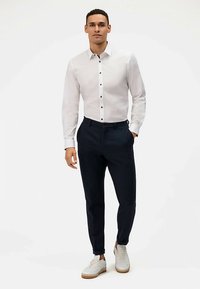 Witte lange mouwen blouse met zwarte knopen, gecombineerd met donkere broek en witte sneakers. Eenvoudig ontwerp, getailleerde pasvorm en minimale textuur.