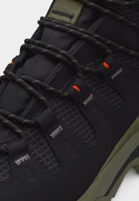 Μαύρο αθλητικό παπούτσι με πλαϊνές πάνελ με σχέδιο πλέγματος, μαύρα και πράσινα κορδόνια περασμένα από μαύρες θηλιές με πορτοκαλί λεπτομέρειες, και σόλα ελιάς.