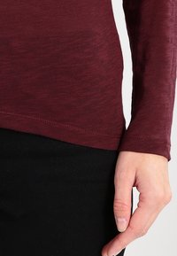 Top burdeos de manga larga con un acabado texturizado. Presenta una forma ajustada y un puño rematado, combinado con una tela negra en la parte inferior.