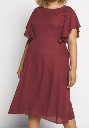 Woman wearing a knee-length burgundy dress with flutter sleeves and a fitted waist, standing against a plain light background.