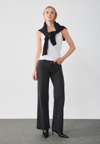 Zwarte hoge taille jeans met wijde pijpen, gecombineerd met een witte tanktop. Een zwarte trui is casual om de nek geknoopt.
