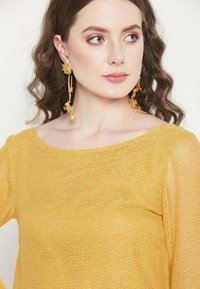Gelber gestrickter Oberteil mit weitem Ausschnitt und langen Ärmeln, der ein lockeres, strukturiertes Gewebe aufweist. Auffällige goldene Blumenohrringe ergänzen das Outfit.