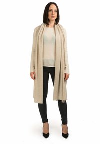 Beige lang cardigan lavet af blødt stof, lagdelt over en lys beige top, parret med mørke skinny jeans og sorte spidse hæle.