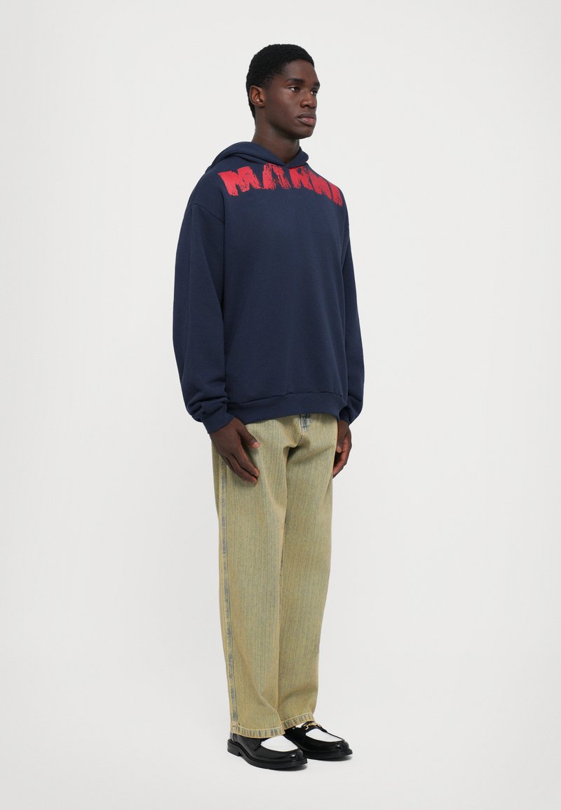 Young man standing wearing navy hoodie with red text, loose light-wash jeans, and black-and-white loafers against a plain background.