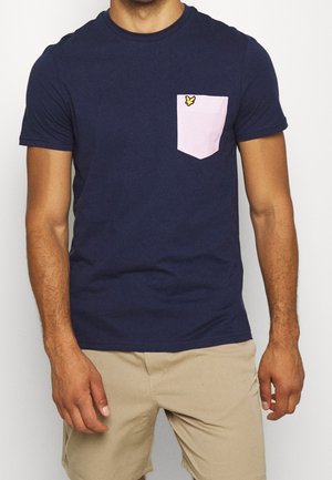 Man wearing navy blue t-shirt with light pink chest pocket and beige shorts, standing with arms relaxed by sides.