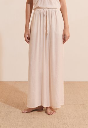 Femme portant un pantalon ample beige clair à jambes larges avec une taille élastique et un cordon, debout pieds nus sur un tapis texturé.