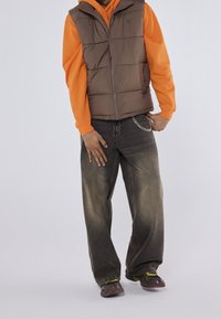 Gilet imbottito marrone con chiusura a zip, indossato sopra una felpa con cappuccio arancione. Jeans larghi sbiaditi con dettaglio a catena. Sneakers con accenti multicolore.