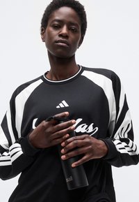 Νέος άνθρωπος με αθλητική στολή Adidas σε ασπρόμαυρους τόνους κρατώντας ένα μαύρο μονωμένο μπουκάλι νερού μπροστά σε απλό λευκό φόντο.