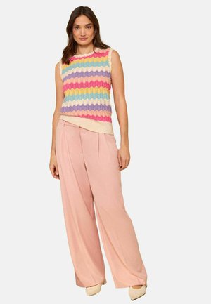 Woman wearing a sleeveless multicolored wavy-striped knit top with light pink wide-leg trousers and beige pointed shoes, standing against white background.