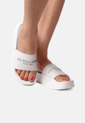 Infradito bianchi con suola texture e ampia cinturino che presenta il testo grigio "U.S. Polo Assn.". Smalto rosso brillante sulle unghie dei piedi.