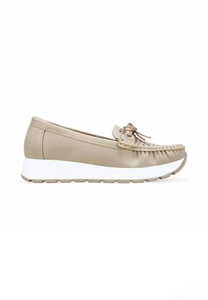 Mocasín beige con puntera cosida, lazo decorativo y suela gruesa de goma blanca, mostrado de perfil sobre un fondo blanco.