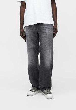 Oseba, oblečena v izprane črne široke kavbojke, belo majico in zelene superge z belimi podplati na enostavni beli podlagi.