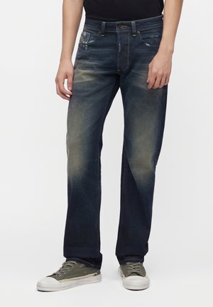 Personne portant un jean droit bleu foncé délavé et des baskets en toile grises, debout devant un fond uni.