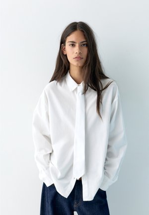 Persoană tânără cu păr lung și închis la culoare, purtând o cămașă albă supradimensionată și o cravată albă, stând cu mâinile în buzunarele unei perechi de blugi de culoare albastru închis, pe un fundal alb.