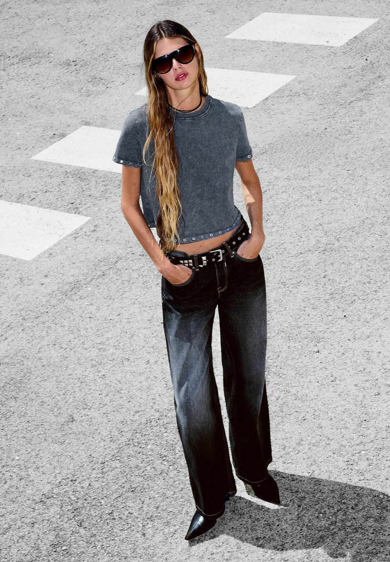Frau mit langen Haaren, die eine dunkle Sonnenbrille, ein ausgewaschenes graues Crop-Top, schwarze weit geschnittene Jeans und spitze schwarze Schuhe trägt und auf einem Zebrastreifen steht.