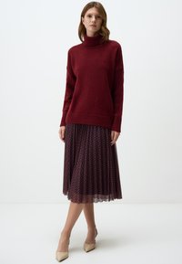 Maglione a collo alto color marsala con lavorazione a maglia testurizzata, abbinato a una gonna plissettata bordeaux con un motivo geometrico. Tacchi a punta beige completano il look.