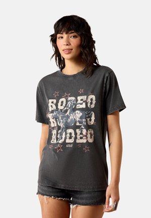 Frau trägt ein ausgewaschenes schwarzes T-Shirt mit „Rodeo“-Grafik mit einem Rodeo-Reiter und Sternen, kombiniert mit schwarzen Jeans-Shorts.