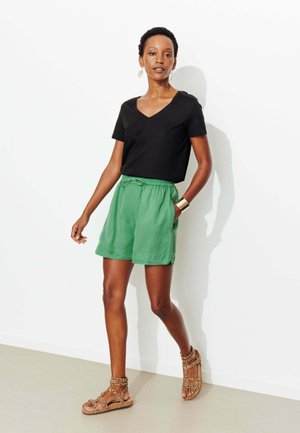 Femme marchant contre un mur blanc, portant un T-shirt noir à col en V, un short vert à taille élastique, des sandales à imprimé léopard, un bracelet manchette en or et des boucles d'oreilles en forme de fleur.