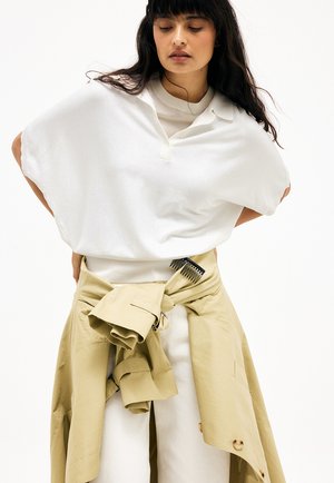 Vrouw met lang donker haar, gekleed in een oversized witte top en een beige jas die op taillehoogte is gestrikt, met een zwarte kam in de jasriem gestoken.