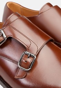 Des chaussures en cuir brun présentent un design à double bride avec des boucles métalliques. Texture lisse avec des détails de coutures visibles et des bouts arrondis.