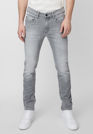 Homme portant un jean slim gris clair et des baskets blanches, debout devant un fond clair uni.