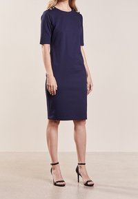Žena v tmavě modrých šatech po kolena s krátkými rukávy a knoflíkovými detaily na ramenou, doplněná černými sandály na vysokém podpatku.
