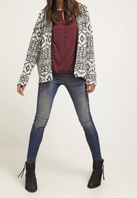 Zwart-wit patroon op een capuchonjack, bordeauxrode top, blauwe strakke spijkerbroek en zwarte enkellaarzen met veters. Gestructureerde materialen en een gelaagd ontwerp.