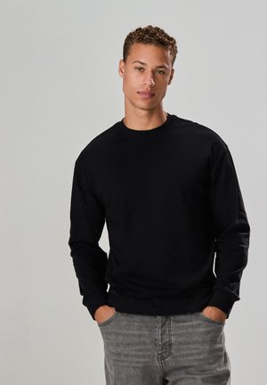 Jeune homme aux cheveux bouclés portant un sweat-shirt noir à manches longues et un jean gris, debout les mains dans les poches devant un fond uni.