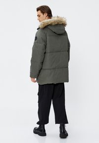 Khakifarbene Puffjacke mit abnehmbarer, mit Pelz gefütterter Kapuze, die ein geräumiges Design und einen strukturierten Stoff aufweist. Trägt dazu locker sitzende schwarze Hosen.