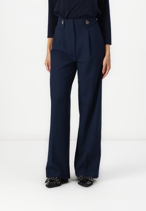 Femme portant un pantalon large taille haute bleu marine avec deux boutons à la taille et des chaussures noires à bout pointu avec des nœuds.