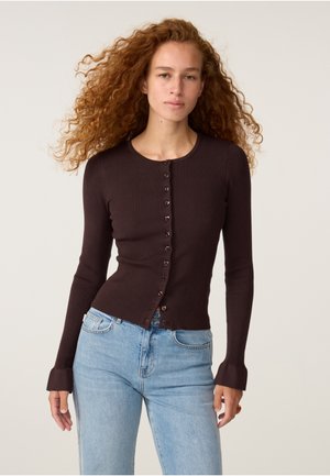 Vrouw met lang krullend haar draagt een aansluitende donkerbruine cardigan met knopen en lichtblauwe spijkerbroek, staand tegen een egale lichte achtergrond.