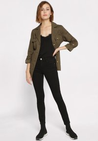 Veste vert olive avec des boutons dorés, débardeur en dentelle noire et jeans skinny noirs associés à des bottines noires. Tissu lisse, design ajusté.