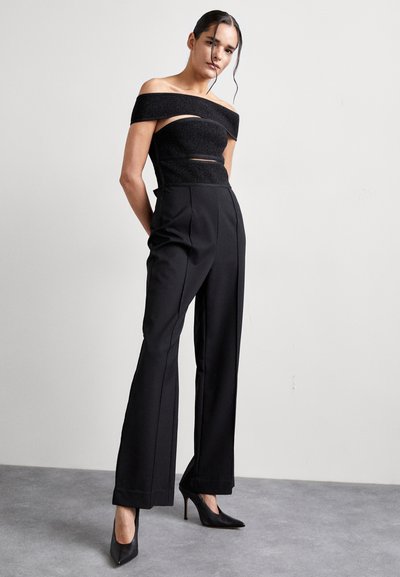 Top negru off-the-shoulder cu un finisaj texturat și detalii decupate, asortat cu pantaloni largi negri cu talie înaltă și tocuri.