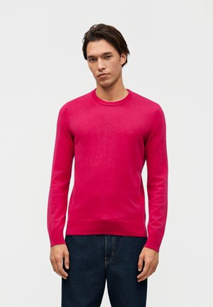 Jeune homme portant un pull ras du cou rose vif et un jean bleu foncé, debout devant un fond blanc uni.