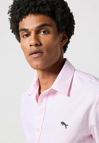 Lichtroze gestreepte shirt met een button-down kraag en een geborduurd donkerblauw logo op de borst. De stof lijkt soepel en ademend te zijn.