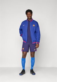 Mand stående i blå og lilla FC Barcelona fodboldudstyr med jakke, shorts, høje sokker og sorte fodboldstøvler mod en ensfarvet baggrund.