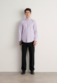 Cămașă lungă cu mânecă și butoni de culoare mov deschis, pantaloni negri și pantofi; poză în picioare; ținând un obiect negru, în fața unui perete simplu.